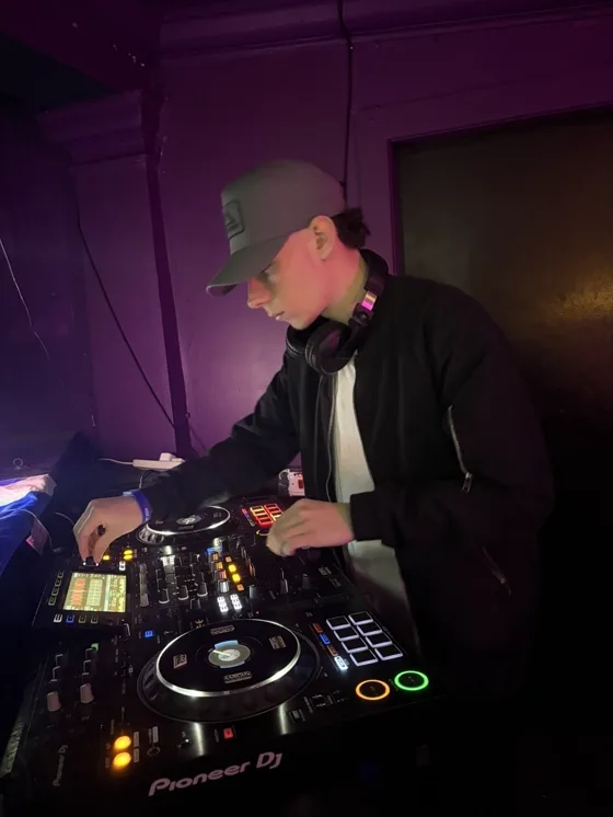 Mixing on the CDJs, headphones round the neck, in a purple-lit booth